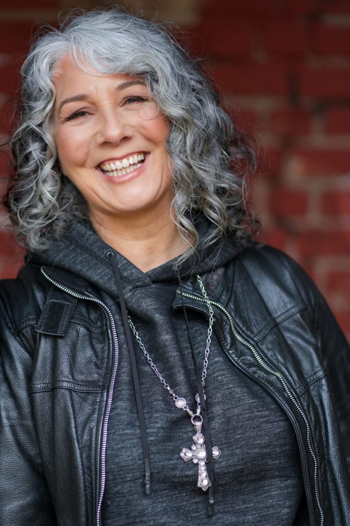Zena Me, a confident woman with grey curls, smiles warmly in a leather jacket, photographed by a London brand photographer.
