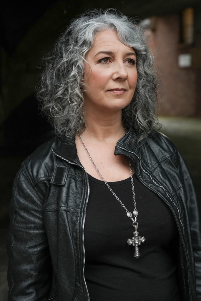 Zena Me, a confident woman with grey curly hair, wears a black leather jacket and a cross necklace. London brand photography.