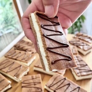 Hand holding a Kinder Milch-Schnitte–style slice with creamy mascarpone filling between two chocolate sponge layers, topped with cocoa and a chocolate drizzle, with more slices on a wooden board in the background.