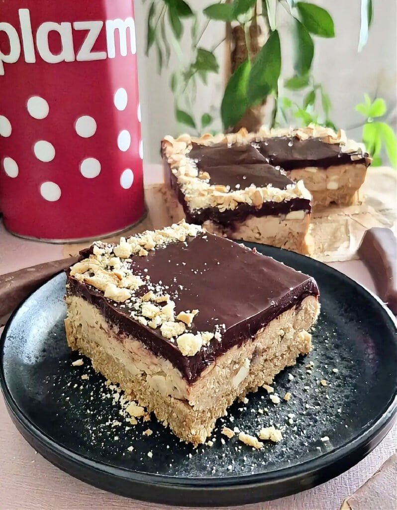 Slice of no-bake chocolate banana cake on a black plate, with another piece in the background, showing the biscuit crust, creamy filling, and chocolate ganache top.