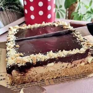 Square chocolate banana no-bake cake with a biscuit crust, creamy middle layer, and glossy chocolate ganache topping, finished with cookie crumbs around the edges.