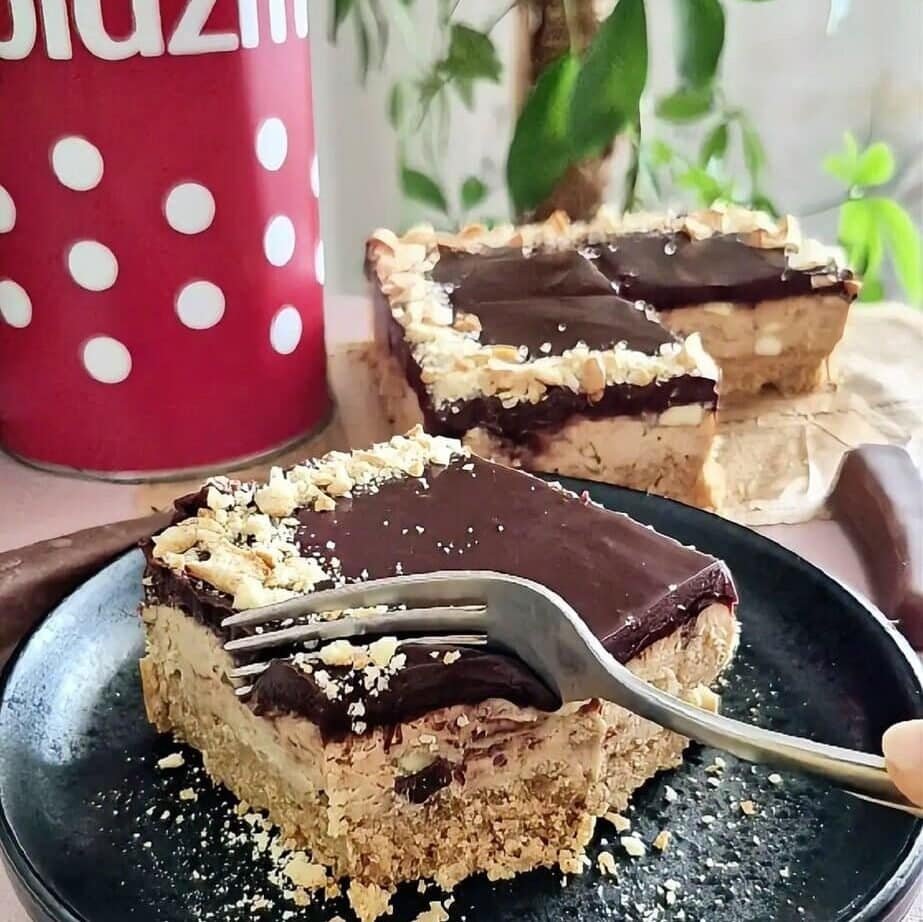 Slice of chocolate banana no-bake cake on a black plate with a fork, showing the biscuit crust, creamy filling, and chocolate ganache top, with extra slices in the background.