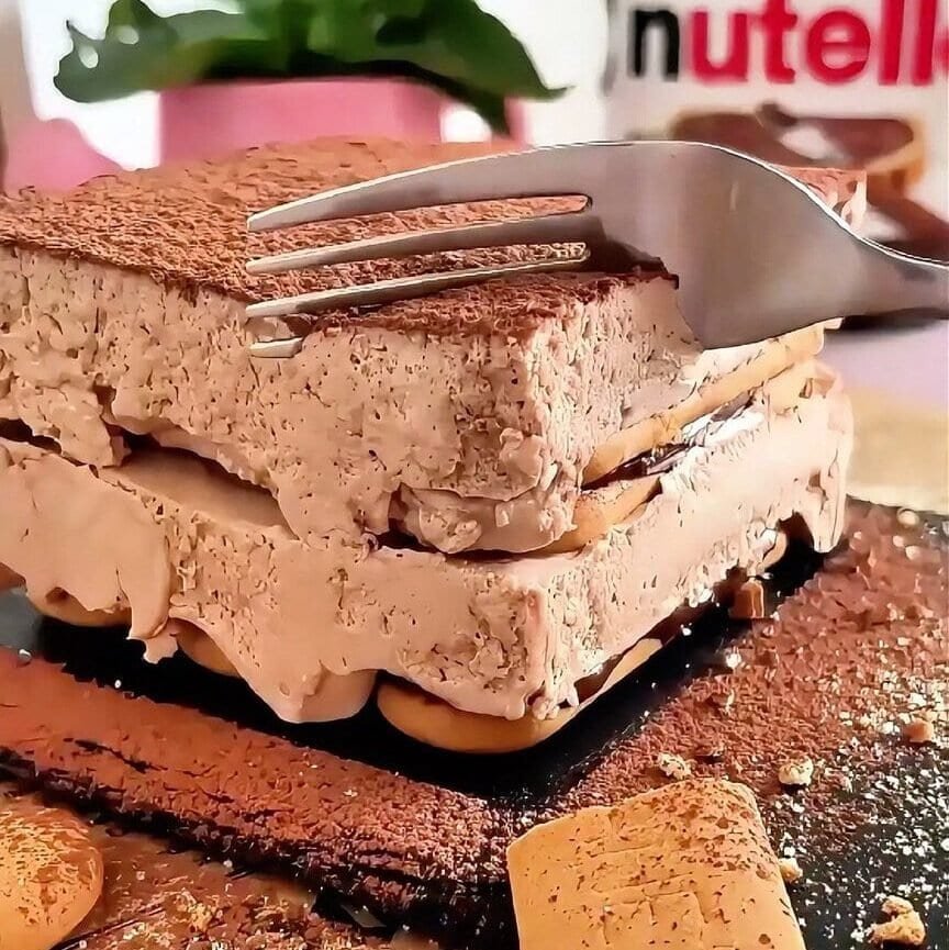 Fork cutting into a slice of mini chocolate biscuit cake layered with Plasmon biscuits and Nutella cream, finished with a cocoa dusting.