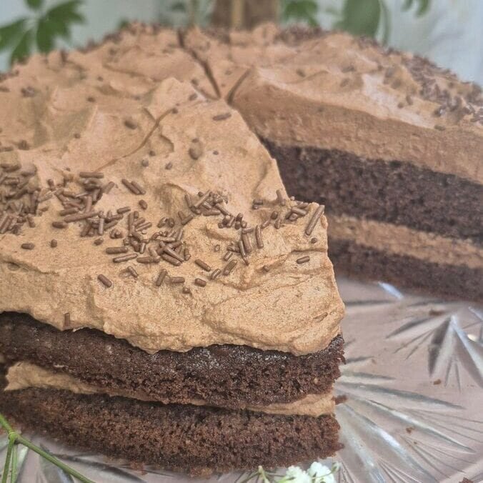 Matilda chocolate layer cake on a glass cake stand with a cocoa-dusted top and a slice cut out, with greenery and small white flowers in the background.