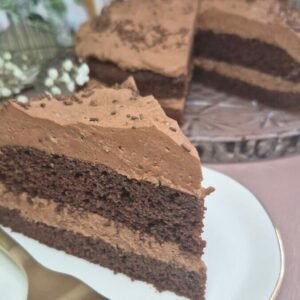 Slice of Matilda chocolate cake with a thick chocolate filling and cocoa-dusted top on a white plate, with more cake pieces in the background.