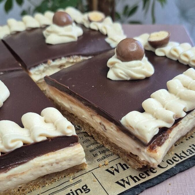 Square no-bake chocolate cheesecake slices with a glossy ganache top and whipped cream dollops, photographed on a purple table with Maltesers nearby.