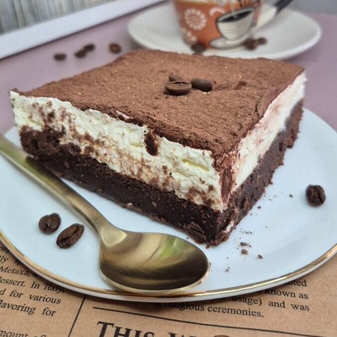 Slice of brownie tiramisu with a coffee-soaked chocolate base, mascarpone cream layer, and cocoa dusting on a plate with a coffee cup in the background.