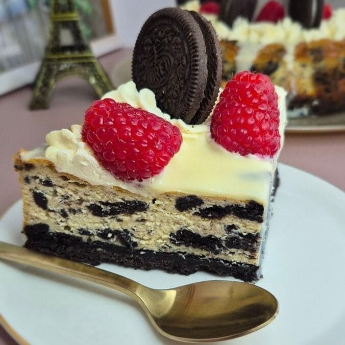 Slice of Oreo cheesecake topped with raspberries and an Oreo cookie on a white plate.
