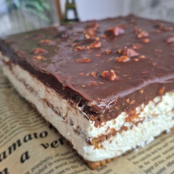 Homemade no-bake Snickers cake with biscuit layers, cream filling, and chocolate topping on a serving surface.