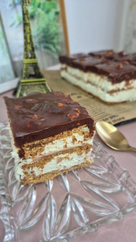 Slice of no-bake Snickers cake with biscuit layers, cream filling, and chocolate topping on a glass plate.