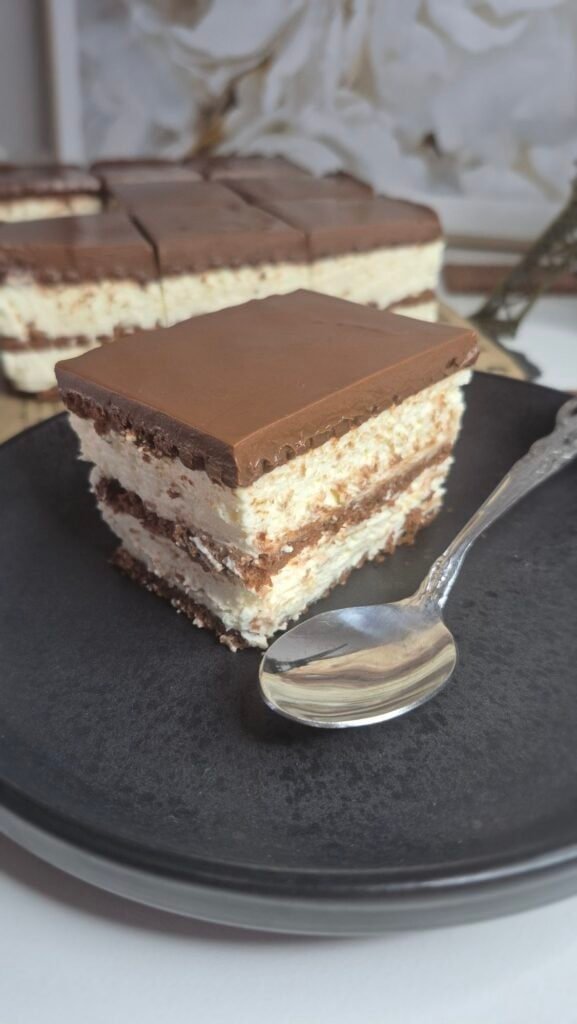 Slice of coffee biscuit cake with mascarpone cream and cocoa topping served on a black plate with a spoon