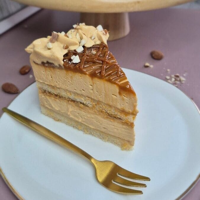 Slice of baked cake with dulce de leche and mascarpone cream on a plate, with the whole layered cake in the background.