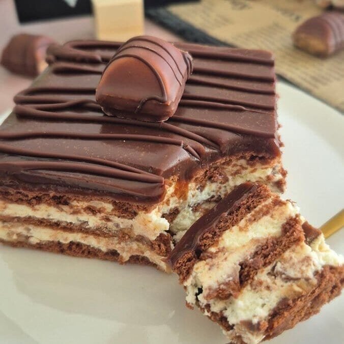 Slice of Kinder Bueno bars showing biscuit and mascarpone layers, topped with chocolate drizzle and Kinder Bueno pieces, served on a white plate.