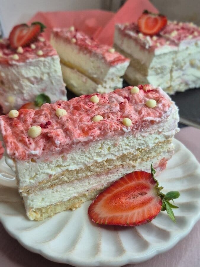 Strawberry tiramisu cake slice on a white plate with mascarpone cream layers, ladyfingers, and fresh strawberry topping, with more slices in the background
