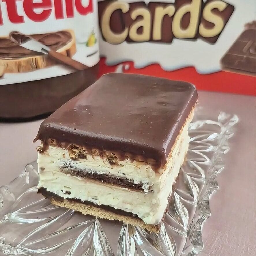 Slice of no-bake Kinder Cards cake with biscuit layers, creamy mascarpone filling, and smooth chocolate ganache, served on a glass plate with Kinder Cards and Nutella in the background.