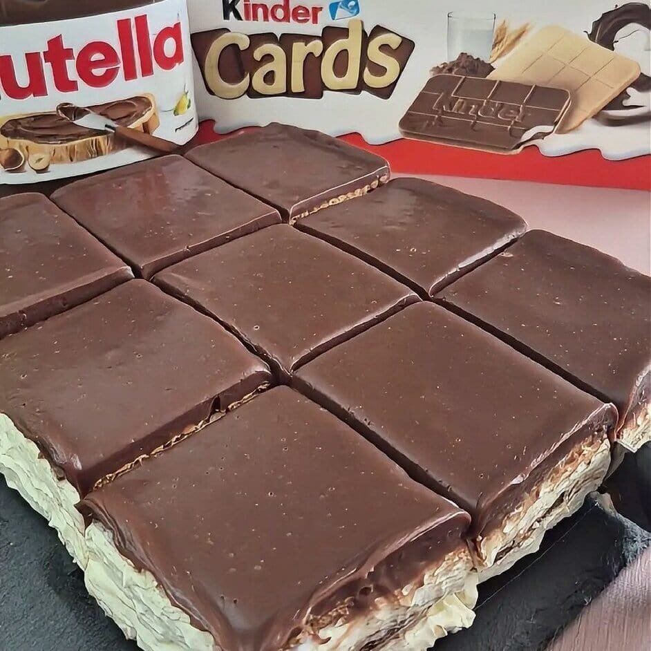 Square no-bake Kinder Cards cake with creamy white filling and smooth chocolate ganache cut into slices, with Kinder Cards and Nutella jars in the background.