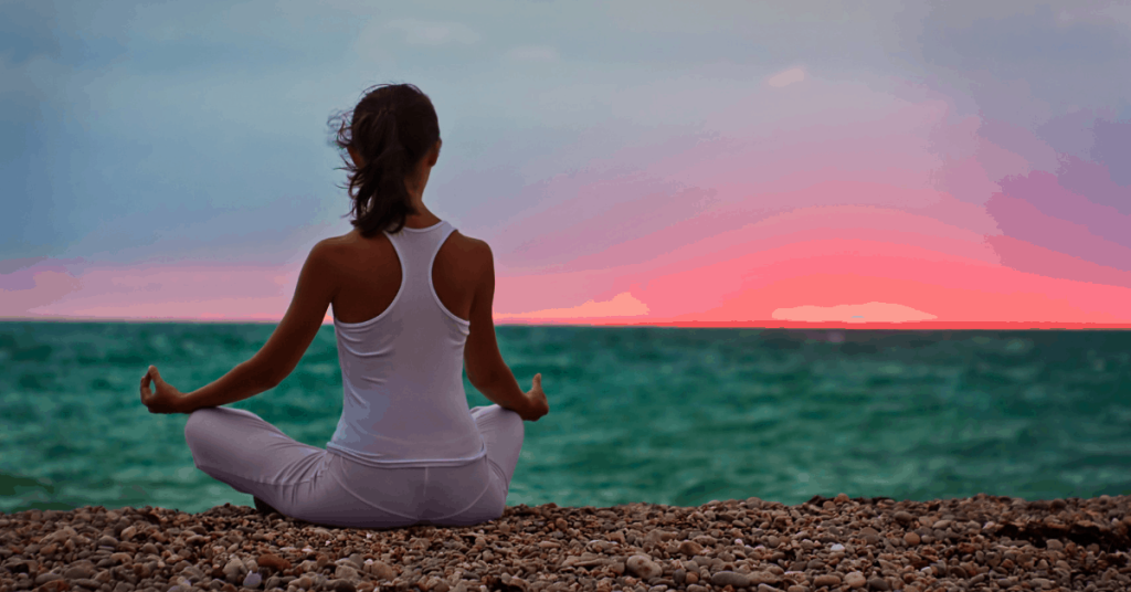young woman meditating and facing body of water with colorful sunset