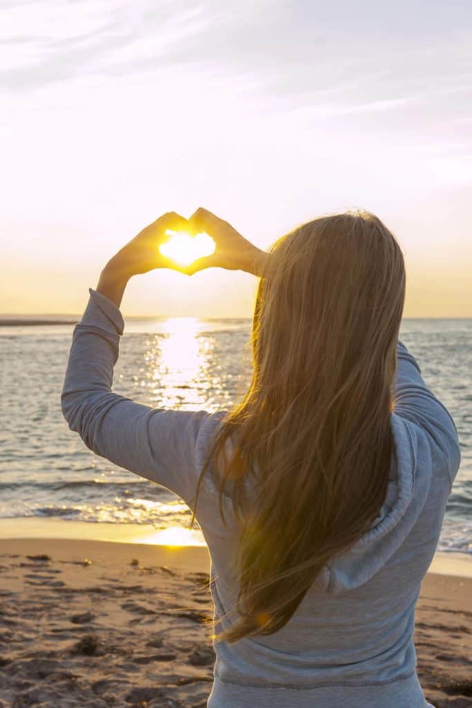 brunette woman on beach facing the water and sunset holding hands in the shape of a heart