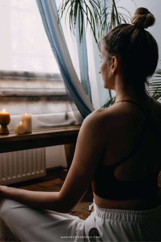 brunette woman with a bun faces window with candles in meditative pose
