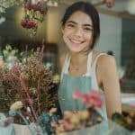 Through window of charming female worker in apron standing among flowers in store smiling at camera