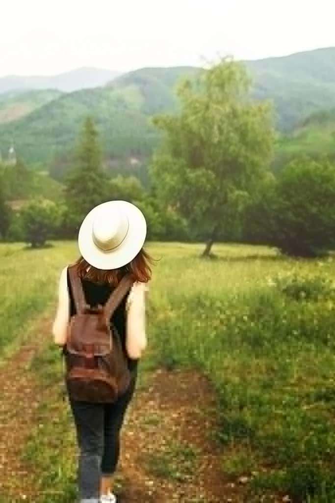 woman in hat and wearing a backpack heads out on a hiking trail in nature with green mountains off in the distance