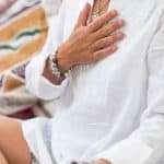 woman practicing Self-Healing Heart Chakra Meditation. Woman sitting in a lotus position with right hand on heart chakra and left palm open in a receiving gesture. Self-Care Practice at Home manifesteveryday.com