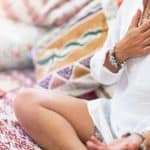 Self-Healing Heart Chakra Meditation. Woman sitting in a lotus position with right hand on heart chakra and left palm open in a receiving gesture. Self-Care Practice at Home manifesteveryday.com