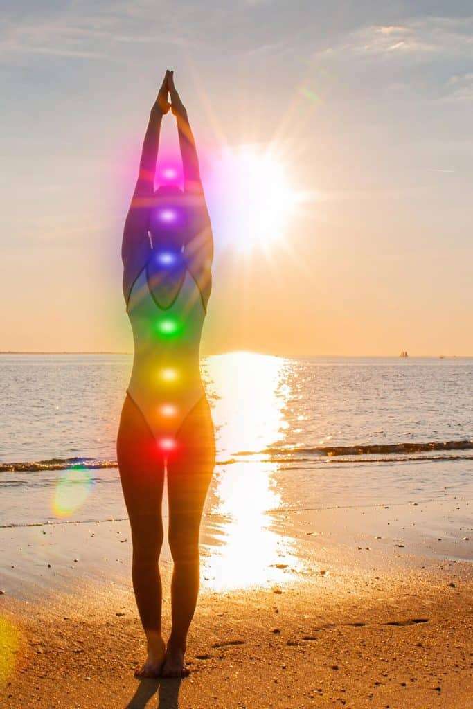 woman standing on beach with arms raised at sunset with chakra colors over her body