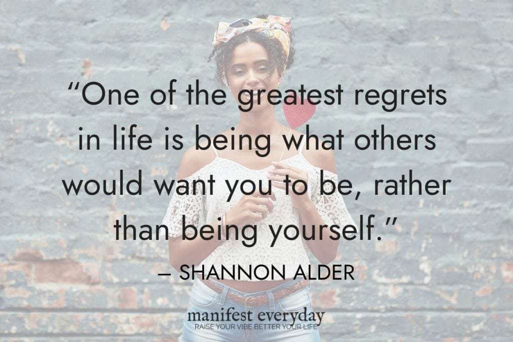 young woman holding heart on stick in front of brick wall with quote text that reads One of the greatest regrets in life is being what others would want you to be, rather than being yourself. — SHANNON ALDER manifesteveryday.com