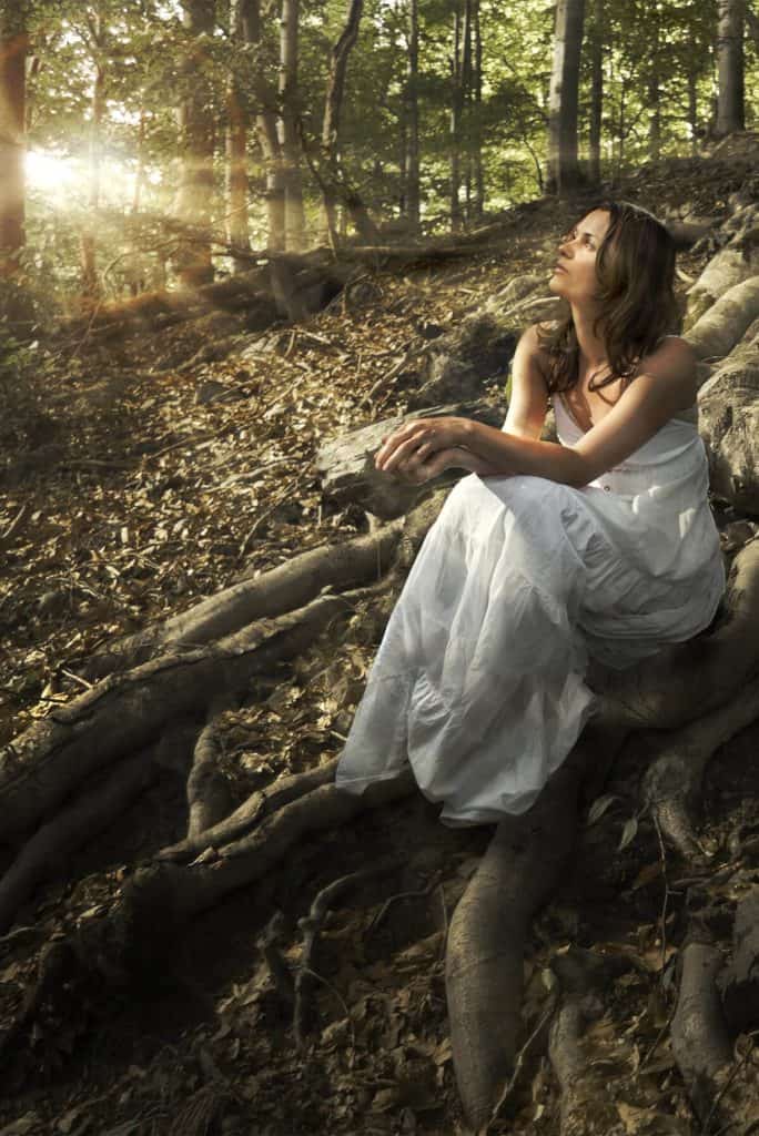 woman in white dress sits on large tree root in the woods gazing at the sky with sunset in the background
