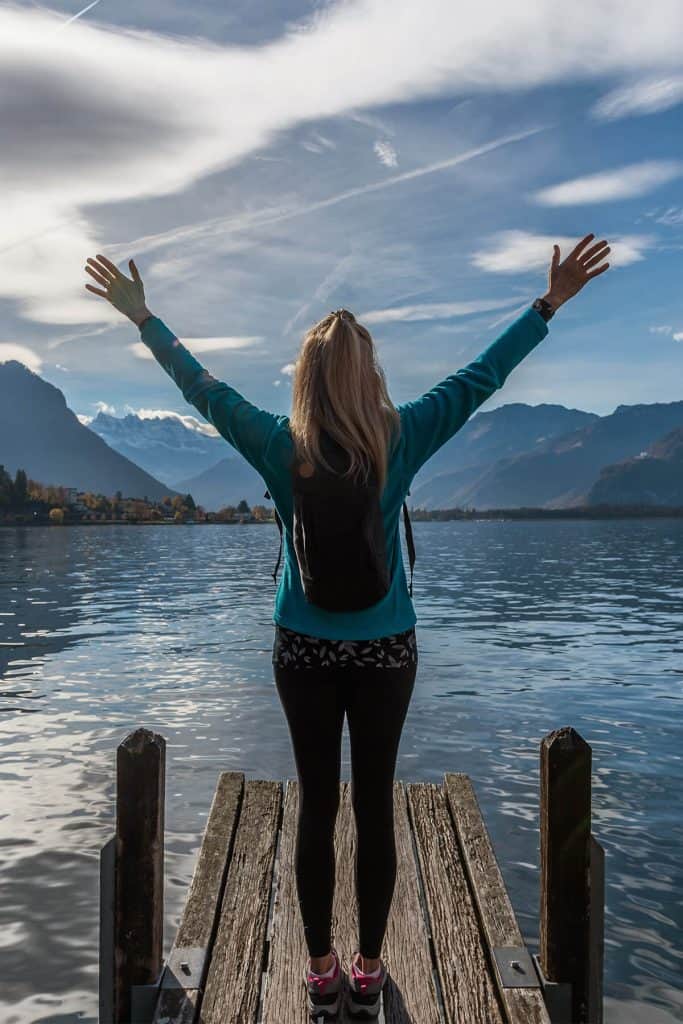 young woman with arms raised stands at the end of a dock looking out at the water, mountains and sky