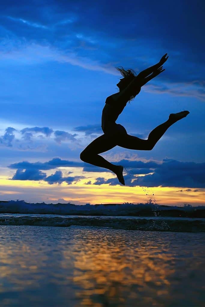 silhouette of woman with arms raised jumping from rock into the water as the sun sets