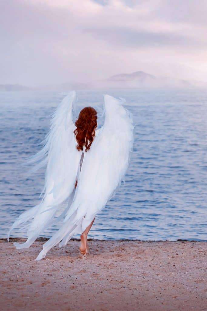 angel with red hair walking towards water with mountains and hazy sky in the background