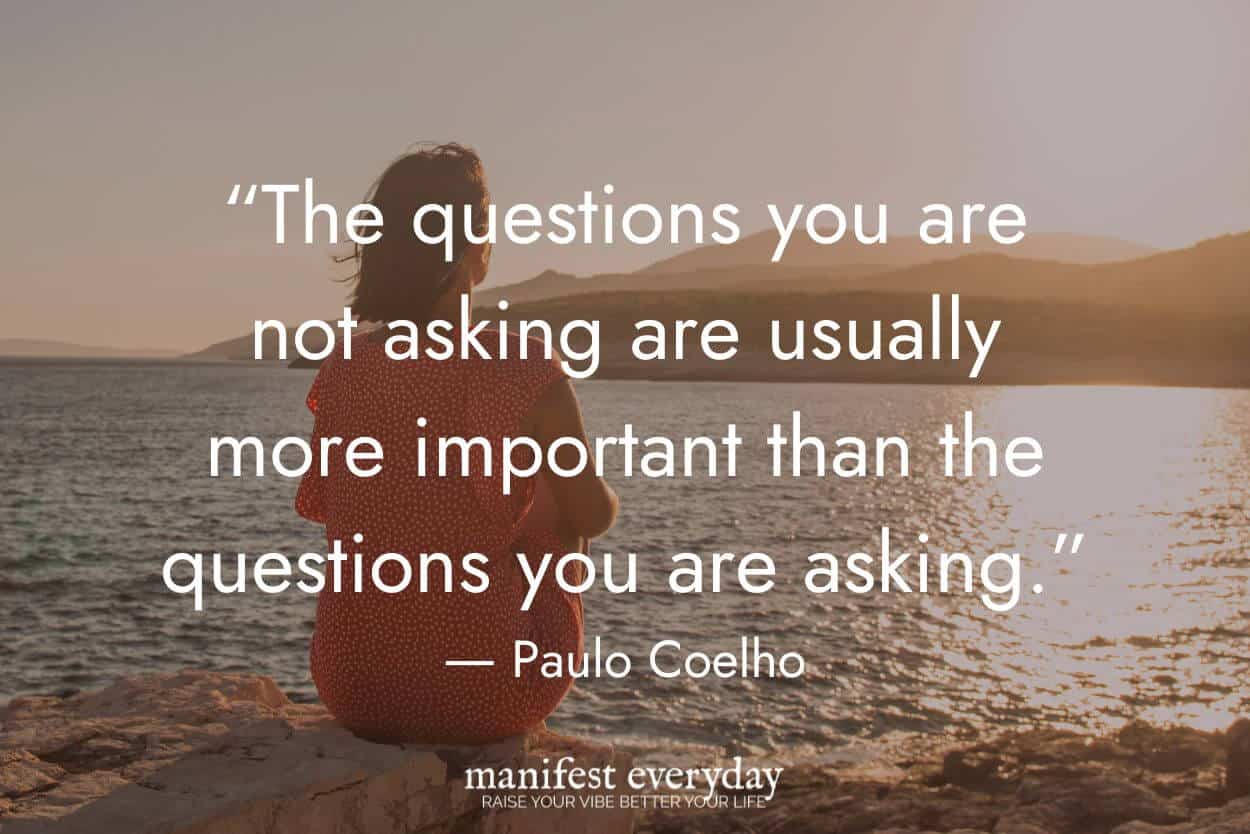woman in red dress sits on rocks by water with mountains in the background at sunset with quote text that reads 'The questions you are not asking are usually more important than the questions you are asking.' - Paulo Coelho manifesteveryday.com
