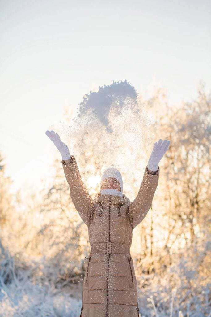woman in coat, mittens and hat throws snow in the air on sunny winter day with frosty trees in the background