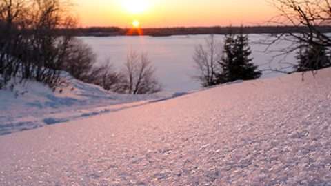 A winter sunset over a snow-covered landscape, with bare trees framing a frozen lake or field. The snow in the foreground sparkles with a pink-orange glow from the setting sun.