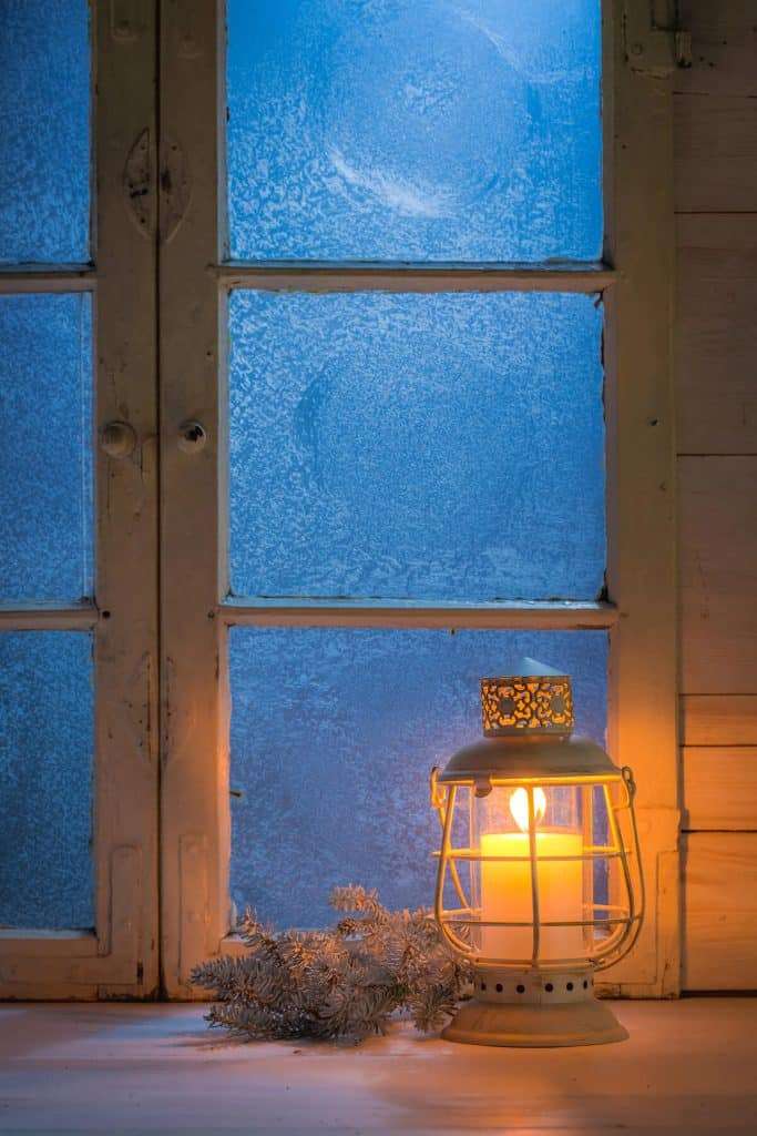 Glowing lantern with candle on winter windowsill with frosted glass panes and evergreen branch, creating cozy hygge atmosphere against blue twilight