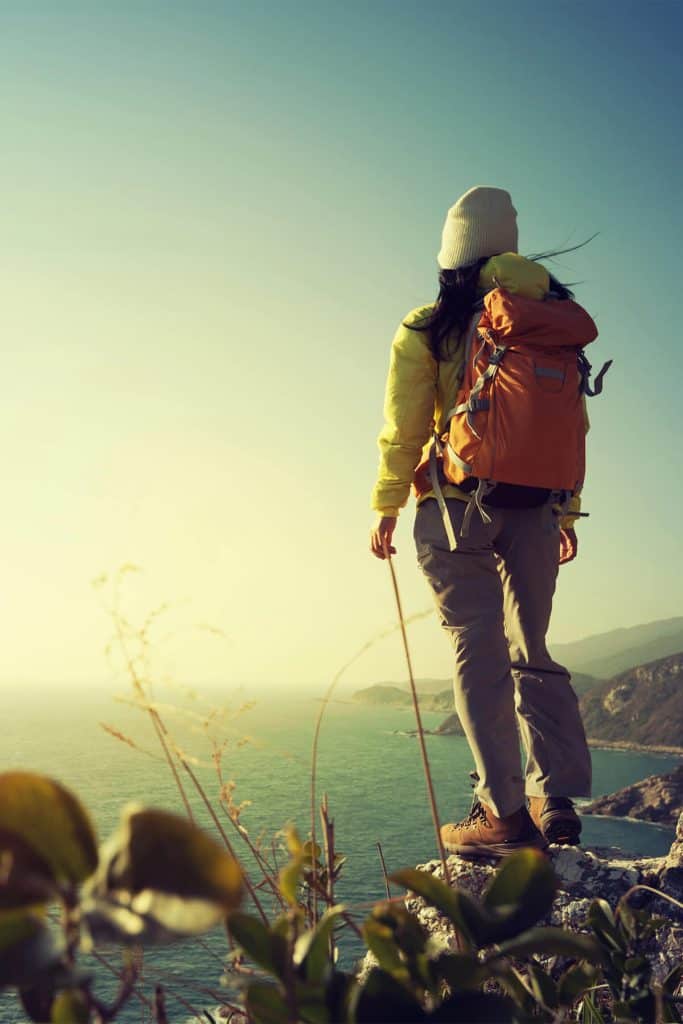 Person in yellow jacket and orange backpack standing on coastal cliff edge overlooking ocean at sunrise, representing personal growth and new beginnings manifesteveryday.com