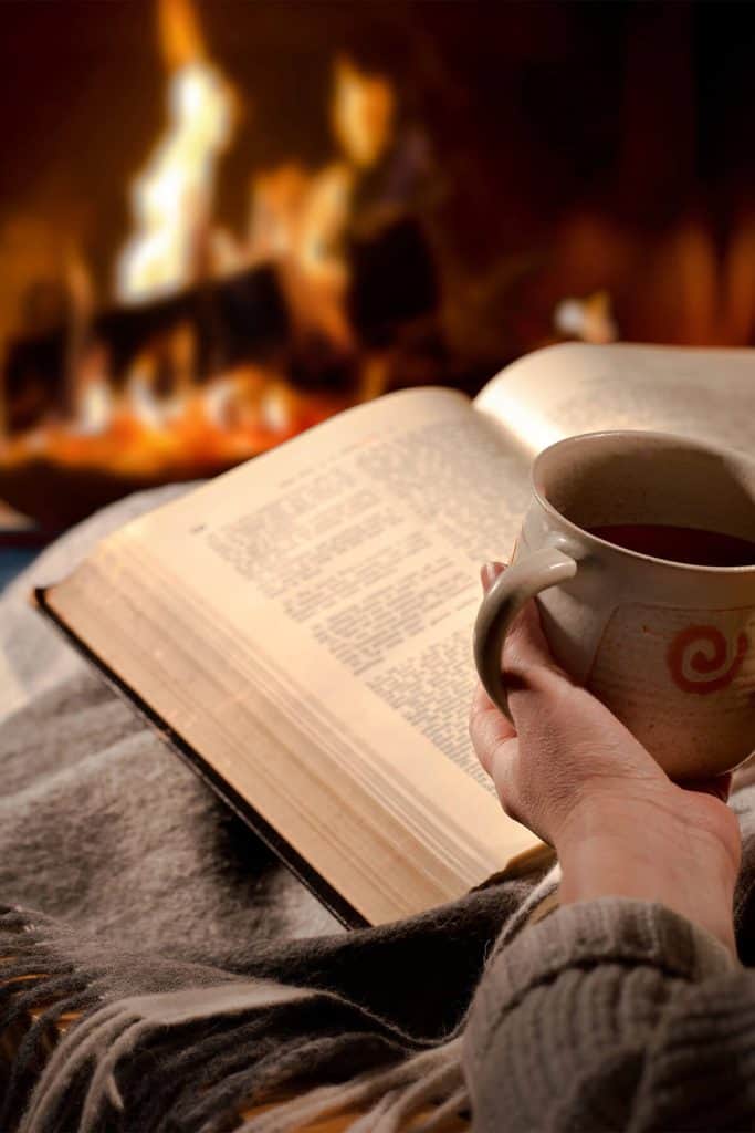 A cozy winter scene with a hand holding a ceramic mug of tea beside an open book, with a warm fireplace glowing in the background. The image captures the essence of slow living and peaceful winter moments.