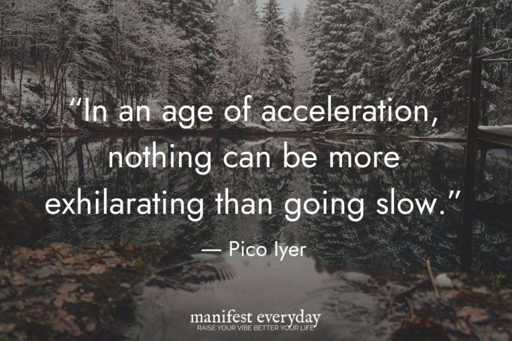 A beautiful winter scene of a perfectly still lake with snow covered trees reflecting in the water with quote text that reads “In an age of acceleration, nothing can be more exhilarating than going slow.” - Pico Iyer manifesteveryday.com