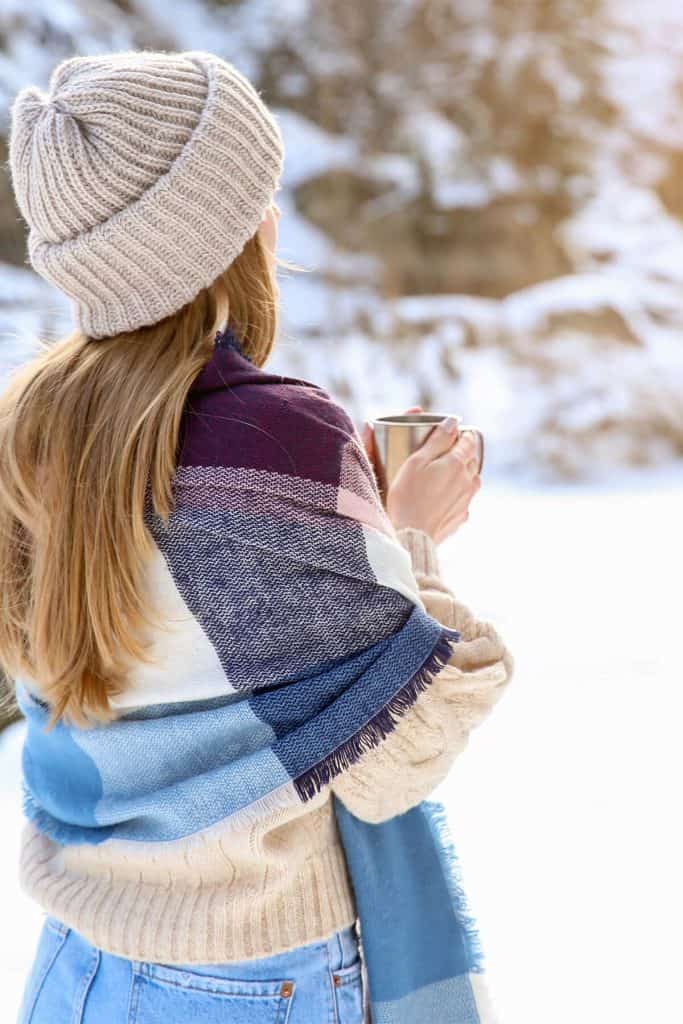 a young woman with hat and shawl holds a mug and looks off into the winter landscape
