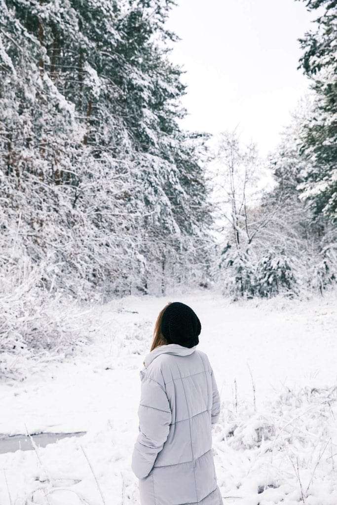 A woman wearing a gray winter coat stands alone on a snowy path in the woods, surrounded by bare trees.
