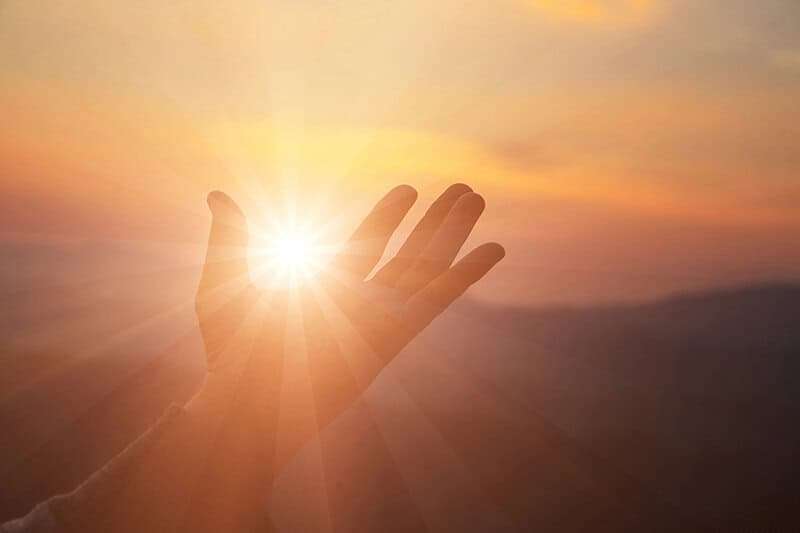 Open hand reaching toward bright sunlight representing manifestation prayer energy and divine connection