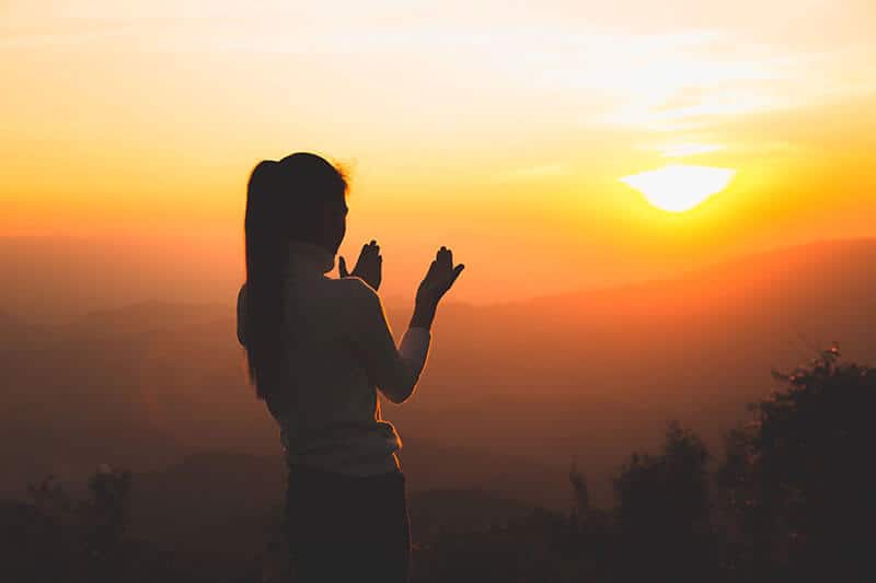 Woman in prayer position silhouetted against golden sunset sky for universe manifestation prayer practice