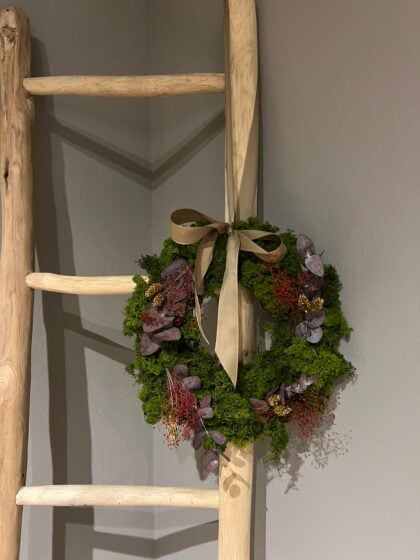 Corona de Navidad con follaje verde, flores moradas y lazo de satén beige, decorativa y elegante.
