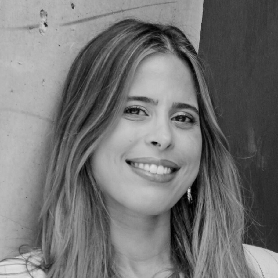 A person with long hair leans against a wall, smiling like a marketing butler effortlessly closing a deal. The image is in black and white.