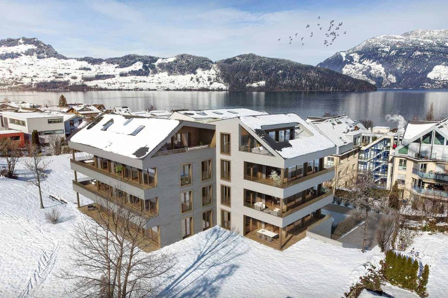 Aerial view of a modern apartment building surrounded by snow with a lake and mountains in the background.