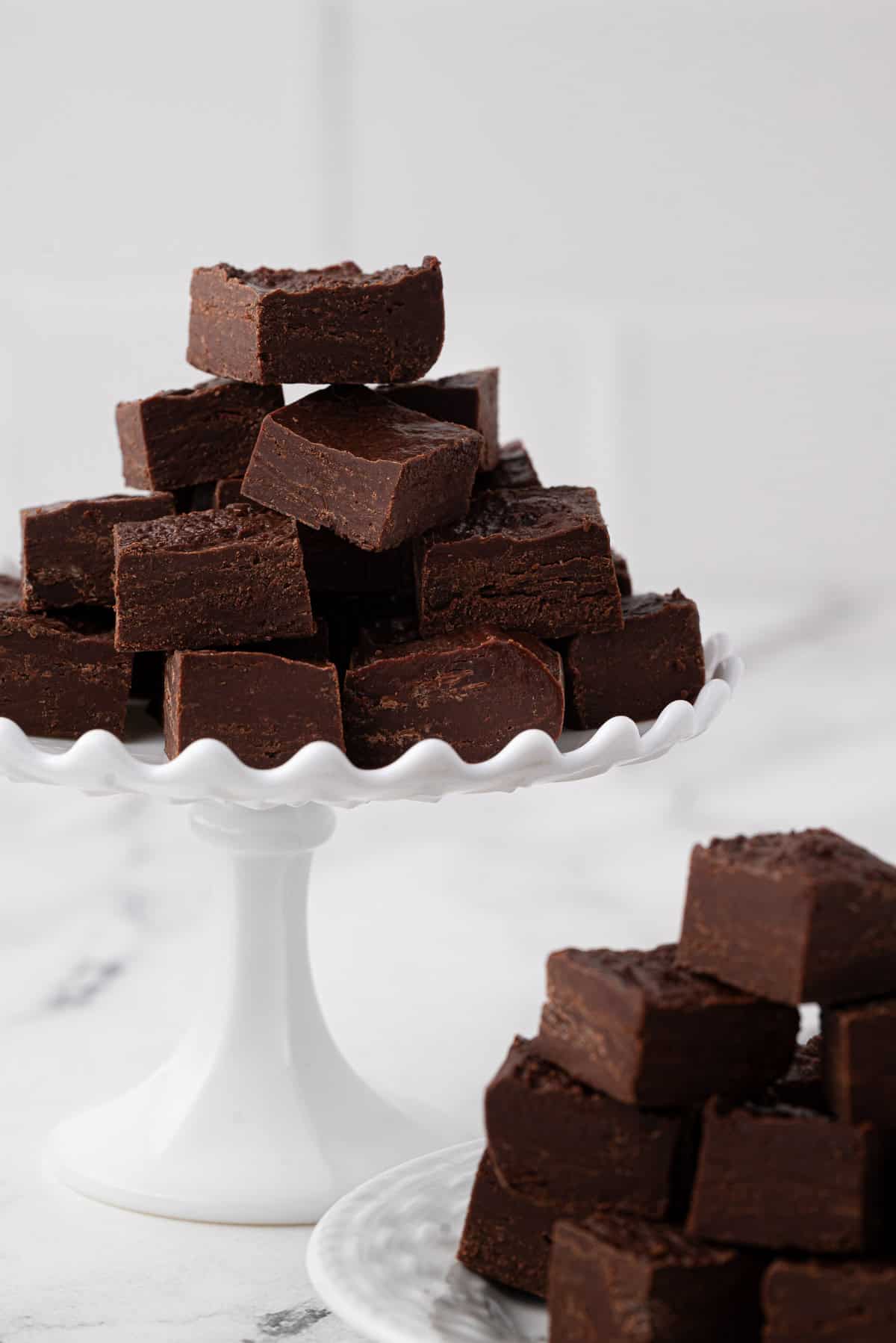 white cake stand with chocolate fudge stacked in a triangle shape with more fudge on a white plate next to it.
