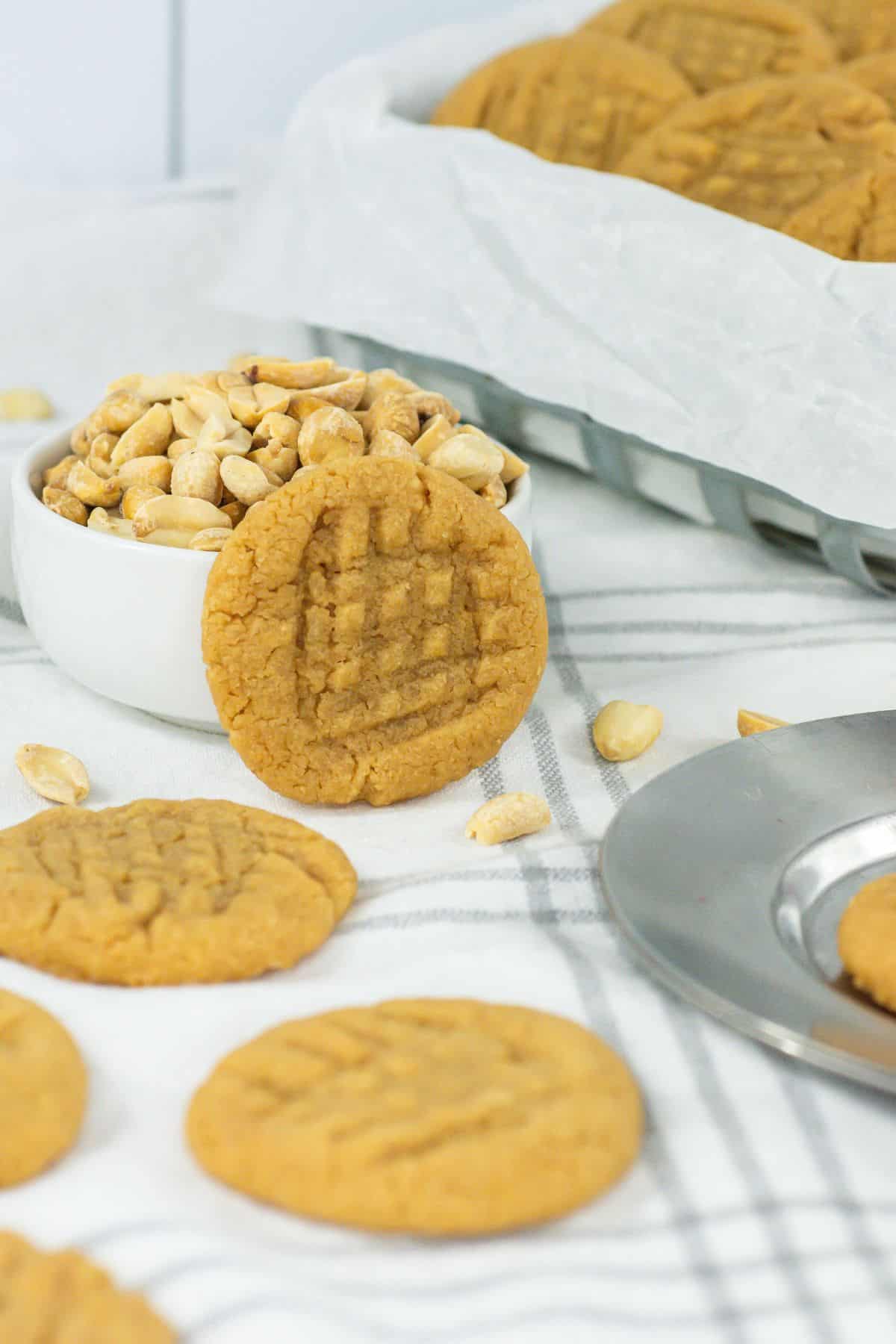 several 3 ingredient peanut butter cookies arranged on a cloth and a cookie leaning against a small white bowl filled with peanuts