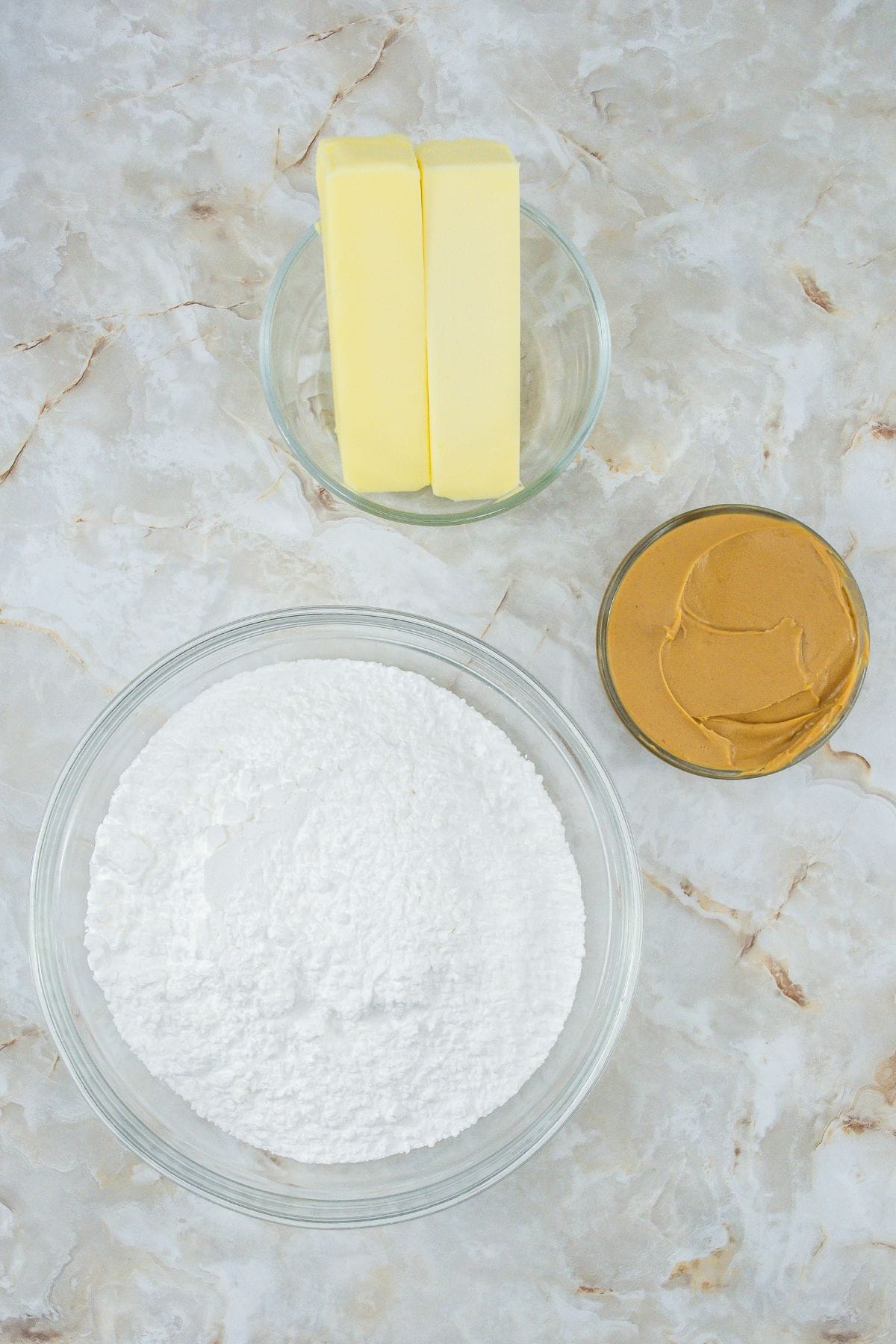three bowls containing each ingredient to make a peanut butter fudge: creamy peanut butter, unsalted butter, and powdered sugar